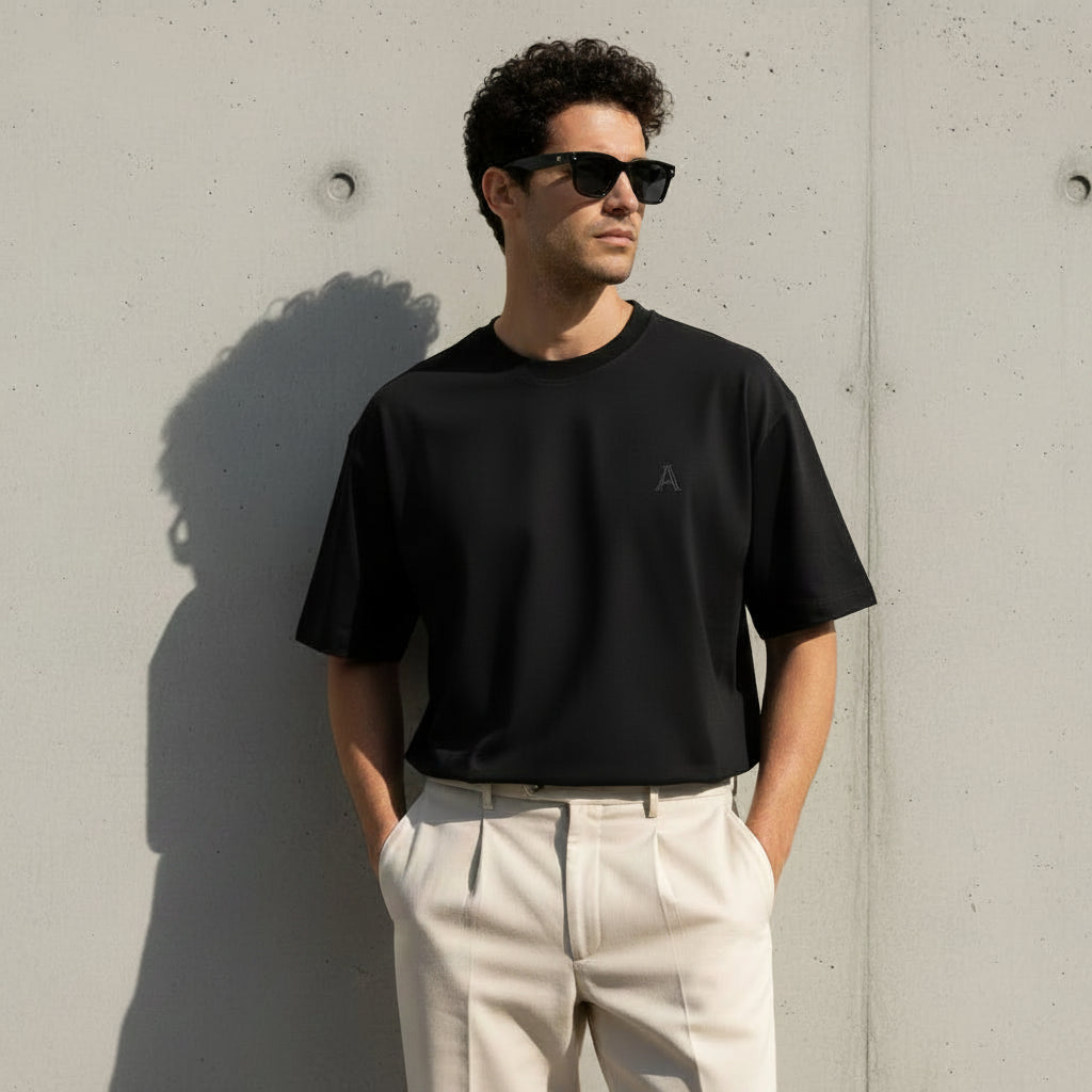 A man wearing an oversized black t-shirt and light beige pants, standing against a textured concrete wall. The image features promotional text on the left reading 'OVERSIZED' with the tagline 'Built for comfort.